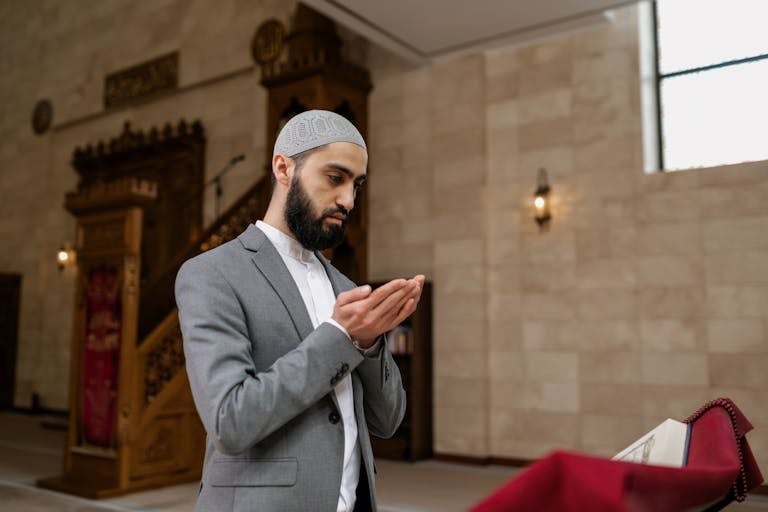 A man in a gray suit, wearing a kufi, prays inside a mosque, hands raised.